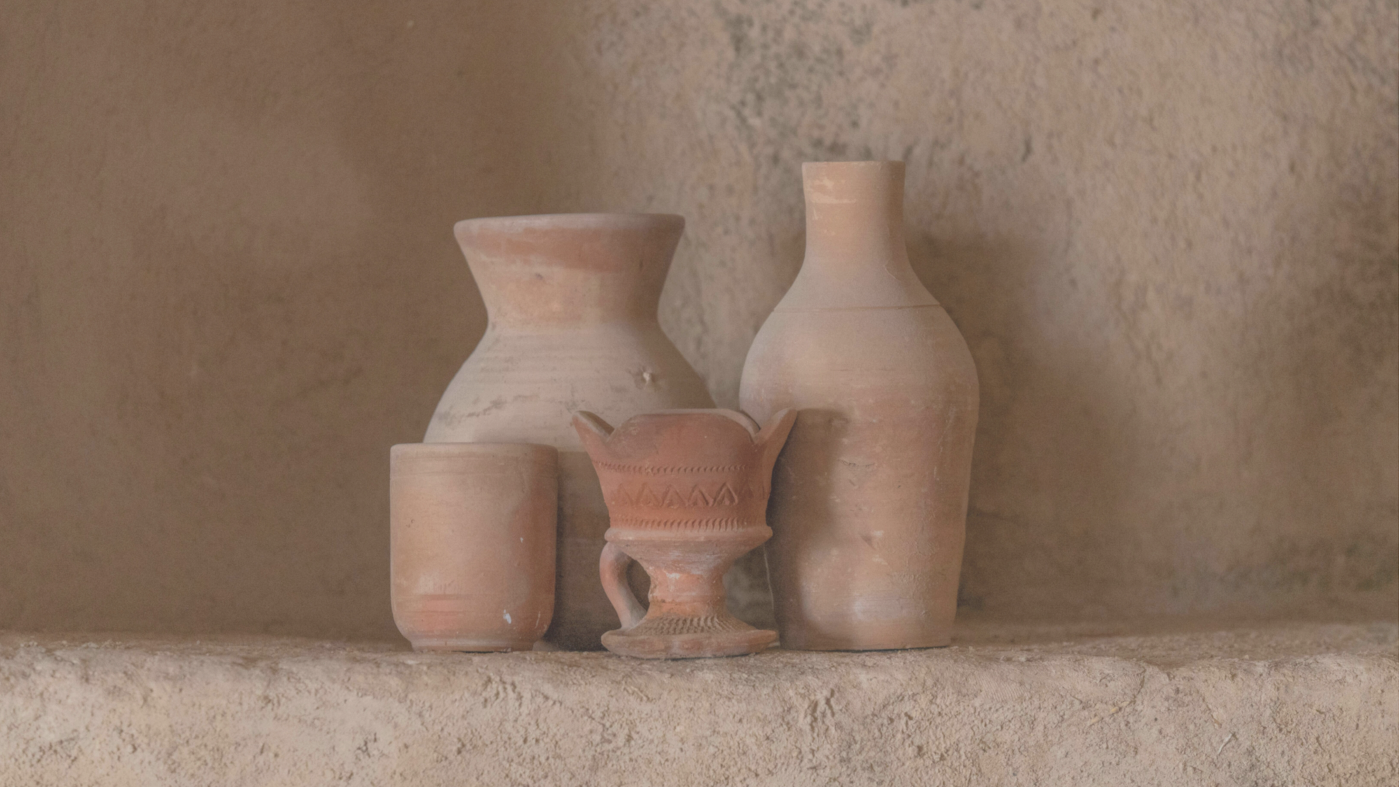 Four simple clay vessels on a quiet shelf against a warm plaster wall.