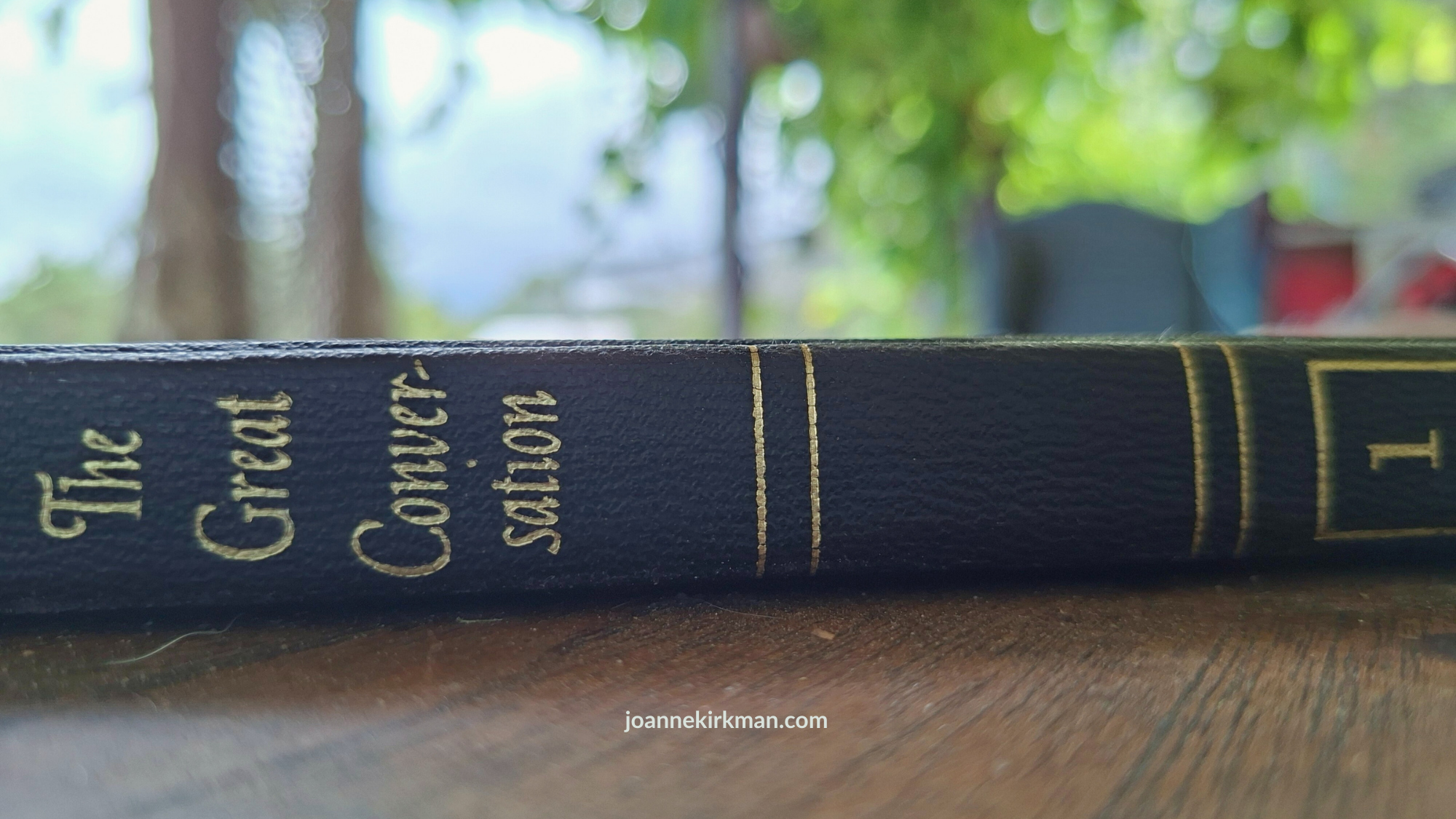 Spine of Mortimer Adler's "The Great Conversation"lying on a wooden table with the garden softly blurred in the background