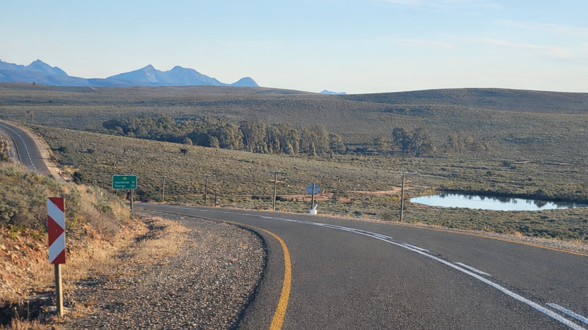 A quiet rural road curves through open Karoo hills beneath a wide blue sky.