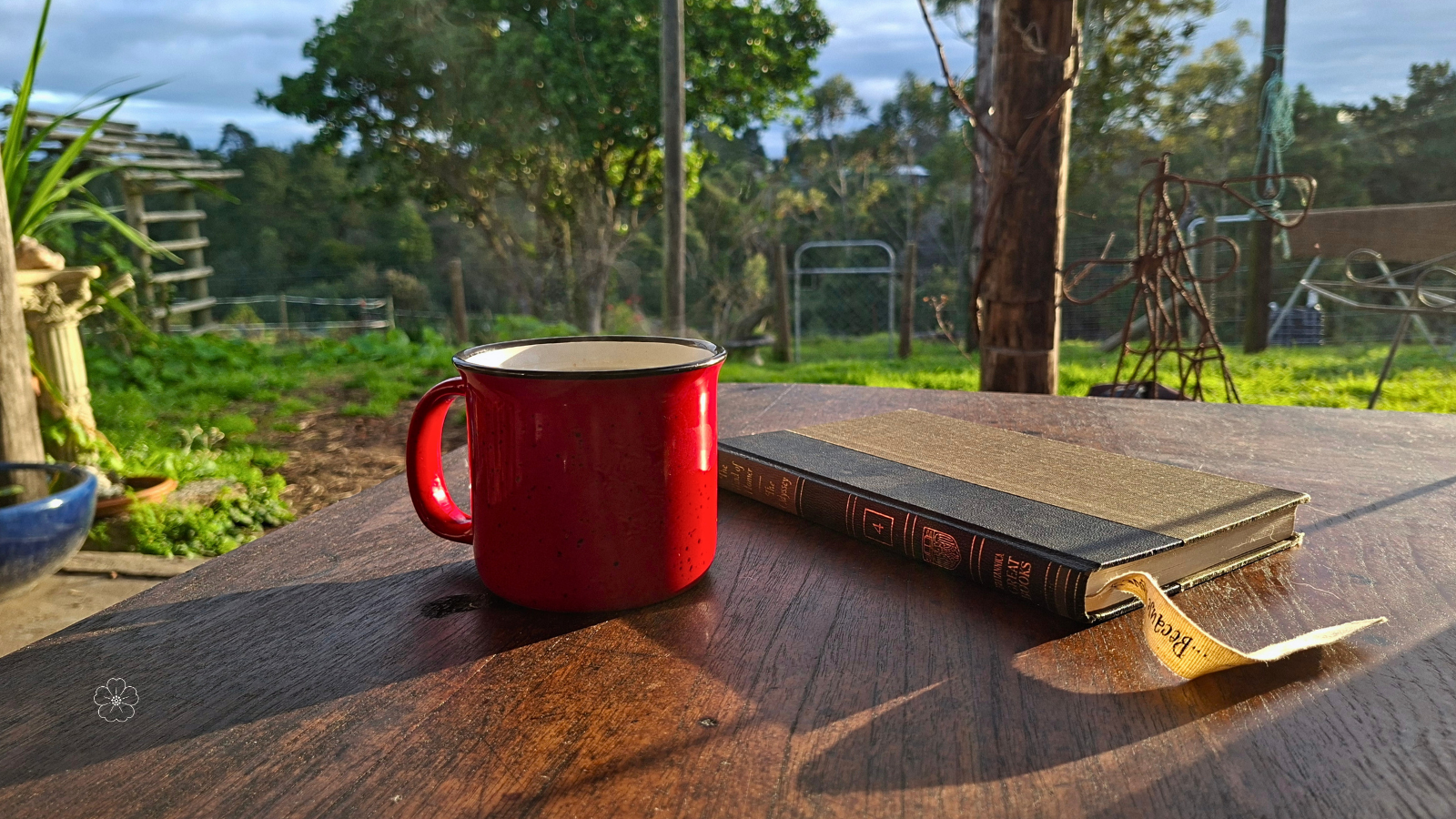 A closed Great Books volume and steaming red enamel mug on a sunlit wooden farm table, with a closed gate and rural landscape beyond.