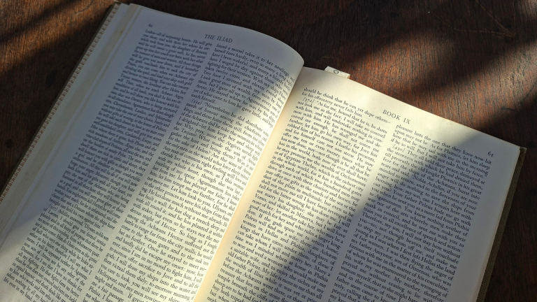An open copy of the Iliad at Book 9 resting on a wooden table in morning light.